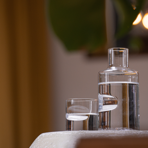 A modern and stylish clear glass barware set up, featuring a stack of cylindrical glass carafes and a small tumbler, arranged on a sleek, light surface. The composition is complemented by soft lighting in the background.