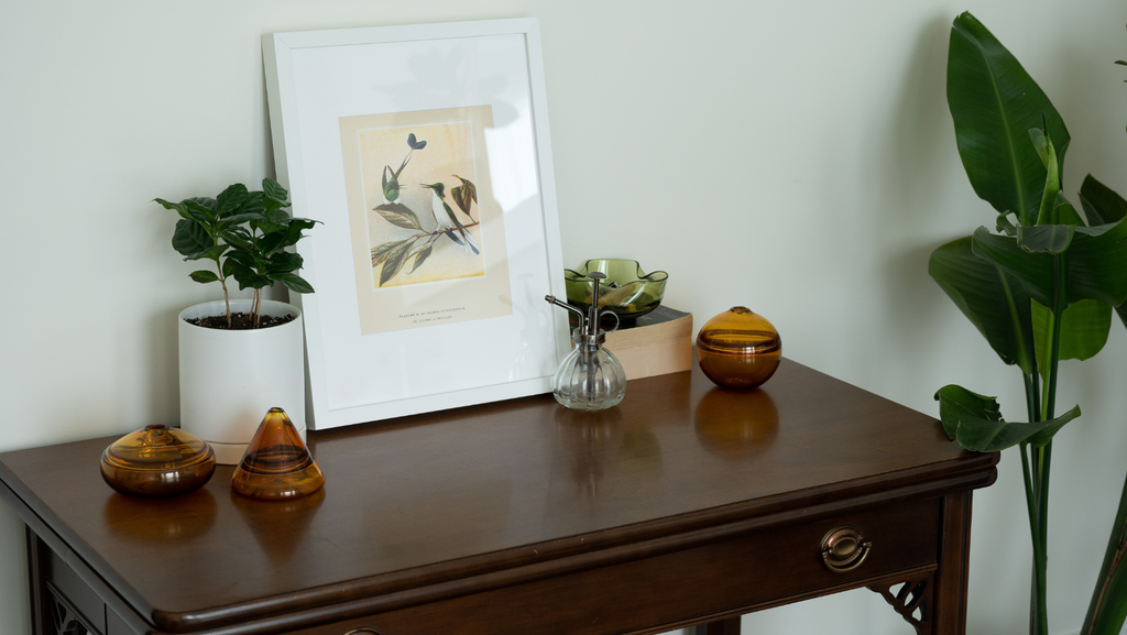 Elegant home decor setup featuring Dougherty Glass pieces on a classic wooden console table. On the left, two amber glass vessels with a reflective finish, and on the right, a clear glass spray bottle and a small amber glass jar