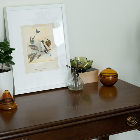Elegant home decor setup featuring Dougherty Glass pieces on a classic wooden console table. On the left, two amber glass vessels with a reflective finish, and on the right, a clear glass spray bottle and a small amber glass jar