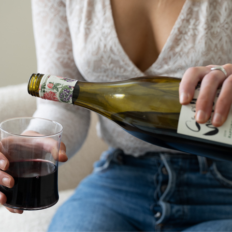 A person in casual attire, focusing on the act of pouring red wine into a glass from a green bottle with a floral label. The setting suggests a relaxed and cozy environment, possibly within a home, capturing an intimate moment of enjoyment.