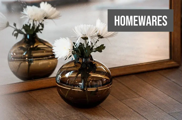 A modern homewares setting featuring a round, reflective brown glass vase on a wooden surface with white flowers, mirrored by its reflection in a large standing mirror, with the word 'HOMEWARES' overlaid in bold, upper-case letters in the upper right corner