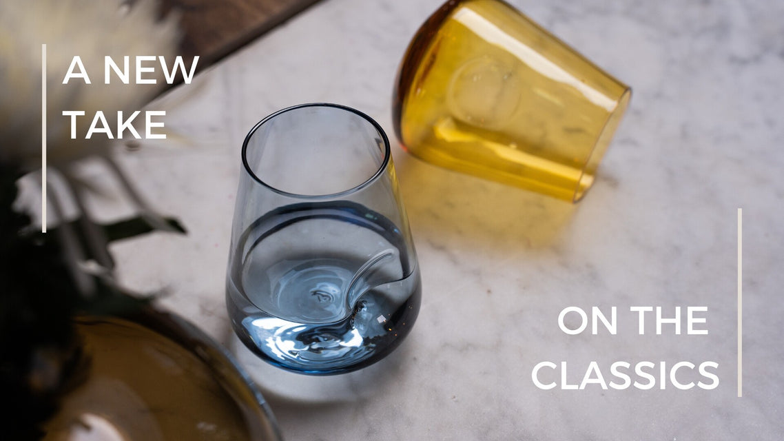 An elegant composition of glassware on a marble surface, showcasing a clear glass with a splash of blue and a tilted amber glass, with the phrase 'A NEW TAKE ON THE CLASSICS' displayed in a modern font, enhancing the contemporary twist on traditional glassware design.