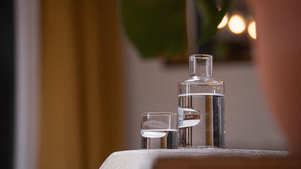 A modern and stylish clear glass barware set up, featuring a stack of cylindrical glass carafes and a small tumbler, arranged on a sleek, light surface. The composition is complemented by soft lighting in the background.