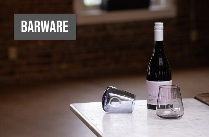 A sophisticated barware setup on a marble countertop featuring one tipped-over clear glass, a standing glass, and a bottle of red wine with a label that reads 'HELLO', all under the bold, upper-case caption 'BARWARE' in the upper left corner against a soft-focused brick wall background.