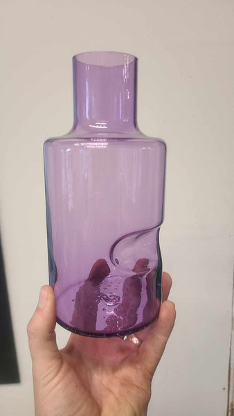 A person holding a lavender-colored glass carafe, showcasing the craftsmanship with a smooth, cylindrical neck that transitions into a wider base with a distinctive curved indent and textured, bubbled bottom. The carafe is partially filled with a liquid, and the holder's fingers are gently curved around its lower part, set against a neutral background.