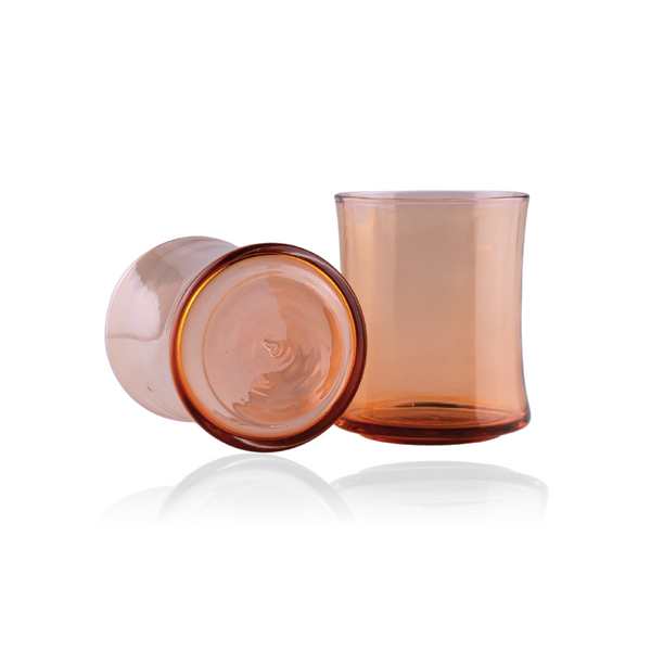 Two rose-tinted glass tumblers are displayed, one resting on its side with its circular base visible, the other standing upright. The glass showcases a subtle gradient of pink, with a darker rim at the base, reflecting on the surface below to enhance the soft, warm glow of the material.