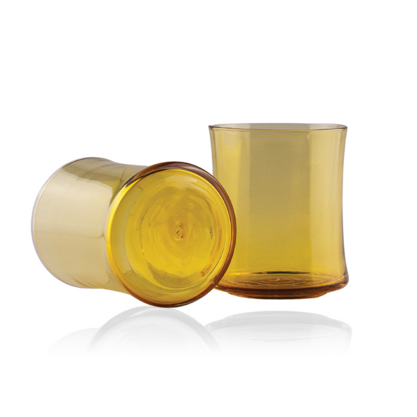 Two amber-colored glass tumblers, one lying on its side and the other standing upright, capture the light to reveal their warm golden hues. Both pieces feature a simple, timeless design, reflected on a polished surface that enhances the glasses' inviting warmth.