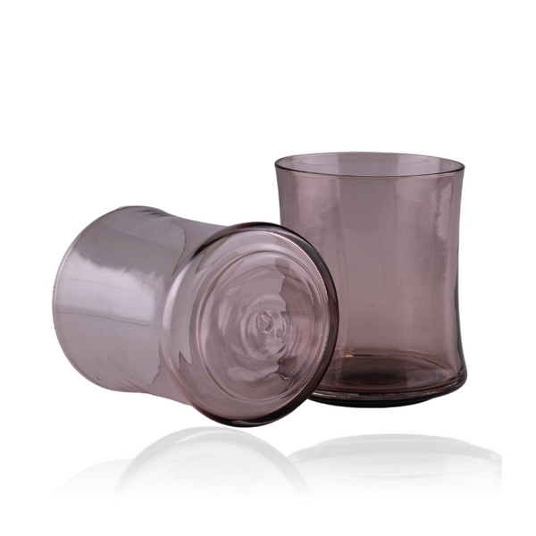 Two smoky mauve glass tumblers are featured here, with one laying on its side showing the base and the other standing upright, reflecting a sense of calm and subtle elegance. The glassware's transparent finish is captured on a reflective surface, enhancing the depth and richness of their hue.