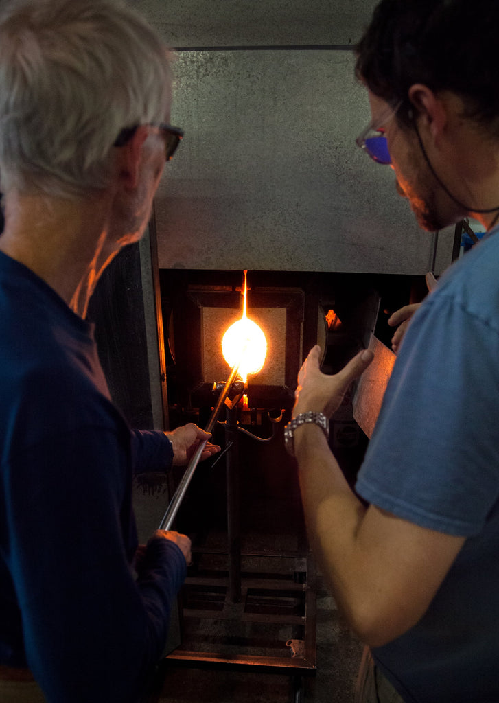 Two glass artists at work in a studio, focusing intently as they shape a glowing molten glass blob at the end of a long metal rod, with the bright interior of a glass furnace visible in the background, highlighting the traditional craft of glassblowing.