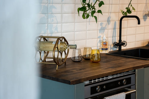 A stylish kitchen counter featuring an antique gold wine rack holding a textured glass bottle, accompanied by two clear and two amber-colored glass tumblers, with a backdrop of white subway tiles and hanging green plants.