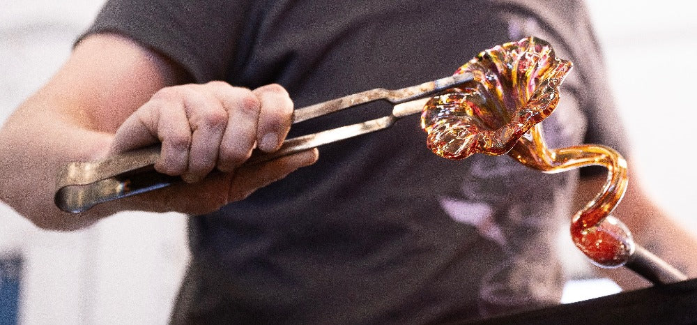 A close-up view capturing the hands of a glass artist using tongs to fine-tune a vibrant, intricately twisted glass sculpture. The piece glows with warm tones of orange and amber, reflecting the artisan's skill and the dynamic process of glassblowing