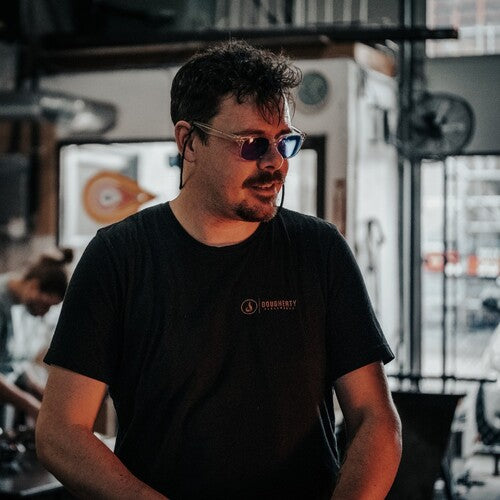 Cameron Dougherty with a confident stance is wearing a casual black t-shirt with a visible logo, paired with blue mirrored sunglasses. He is indoors, with the blurred background of a glassblowing facility.