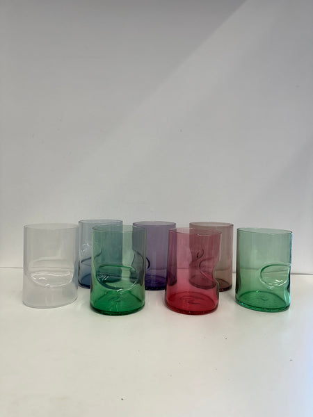 A collection of six handcrafted glass tumblers in a line, each with a unique swirl and color: blush pink, lavender, amethyst, forest green, crimson, and lime. The tumblers feature an indent at the bottom, creating an artistic wave-like effect. They are displayed on a white surface with a soft shadow beneath each, indicating a well-lit environment.