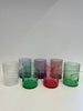 A collection of six handcrafted glass tumblers in a line, each with a unique swirl and color: blush pink, lavender, amethyst, forest green, crimson, and lime. The tumblers feature an indent at the bottom, creating an artistic wave-like effect. They are displayed on a white surface with a soft shadow beneath each, indicating a well-lit environment.