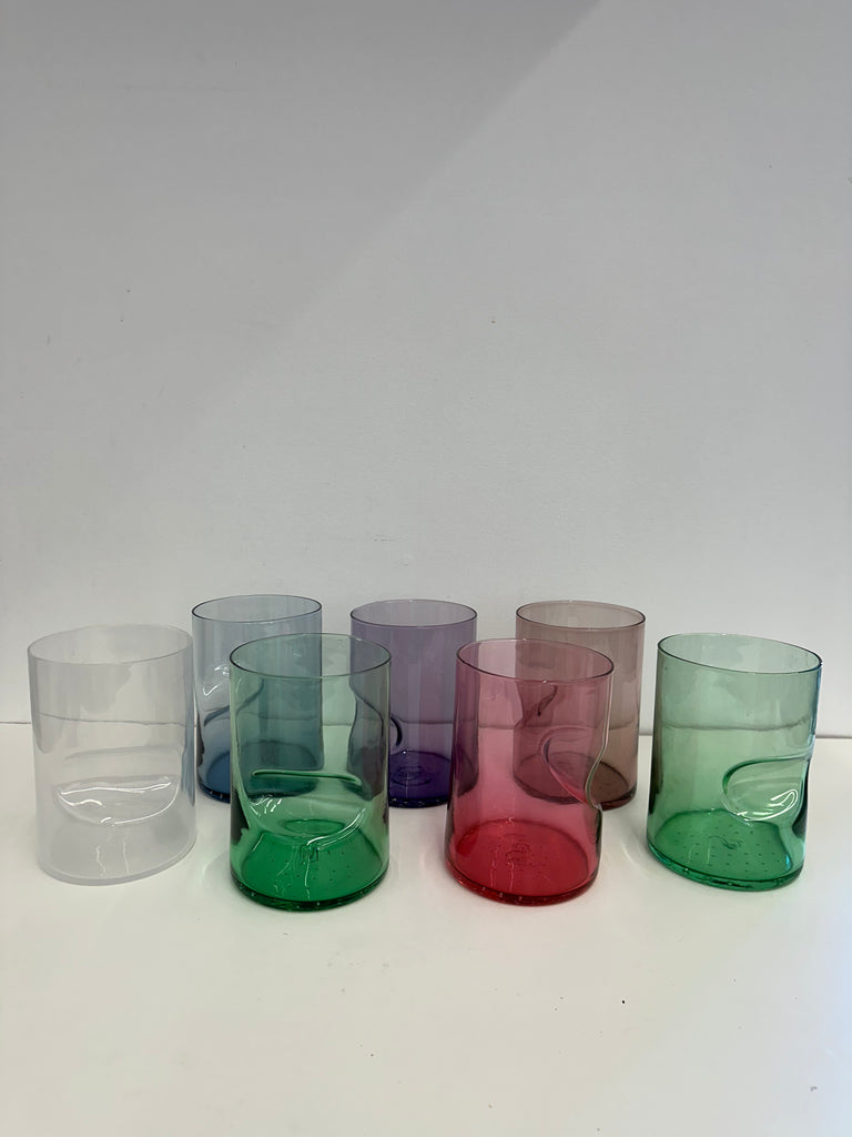 A collection of seven handcrafted glass tumblers in a line, each with a unique swirl and color: blush pink, lavender, amethyst, forest green, crimson, and lime. The tumblers feature an indent at the bottom, creating an artistic wave-like effect. They are displayed on a white surface with a soft shadow beneath each, indicating a well-lit environment.