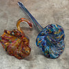 Two handcrafted glass sculptures in the shape of leaves on a textured surface: one with a vibrant mix of red, orange, and yellow tones, and the other showcasing a swirl of blues and purples, each with a gracefully curved stem.