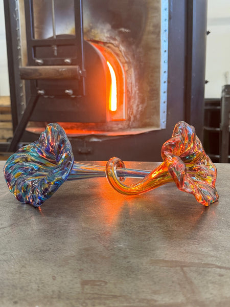 An intricately twisted glass sculpture with vibrant swirls of blue and orange rests on a metal surface in a glassblowing studio, illuminated by the warm glow from the furnace in the background, highlighting the fluid beauty of hand-blown glass art.