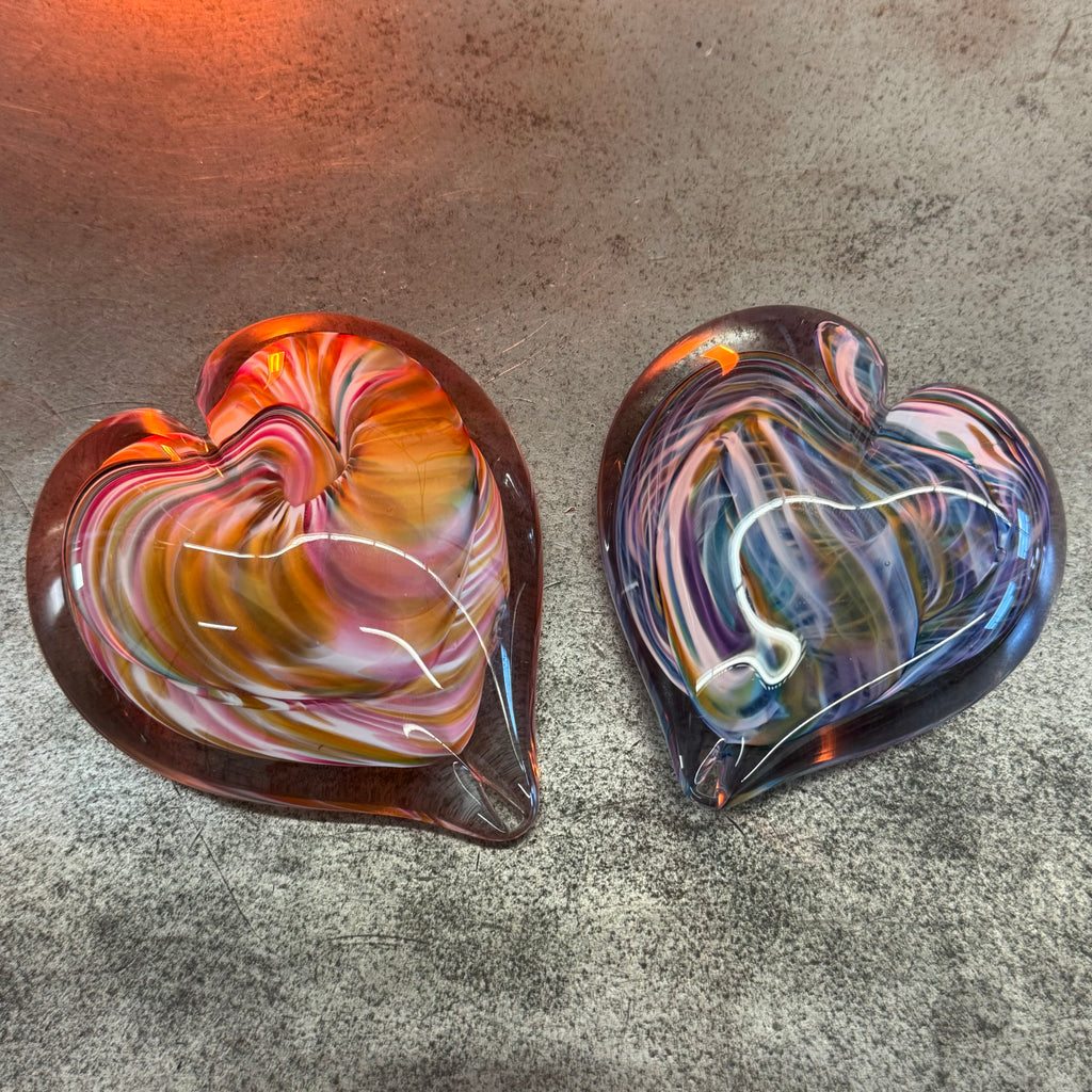Two hand-blown glass heart sculptures resting on a textured surface, one with swirling hues of amber and red, the other in shades of blue and purple, showcasing the artistry of glassmaking with their intricate, colorful patterns and glossy finish.