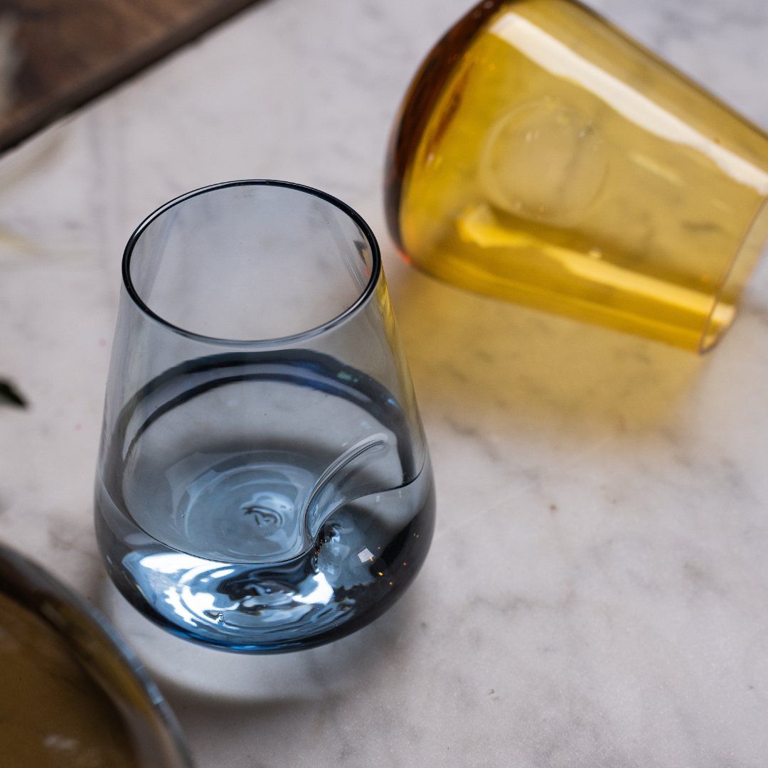 A stemless wine glass with a rounded base and distinctive thumb indent, showcased against a reflective surface with a black backdrop to enhance the glass's warm hues.