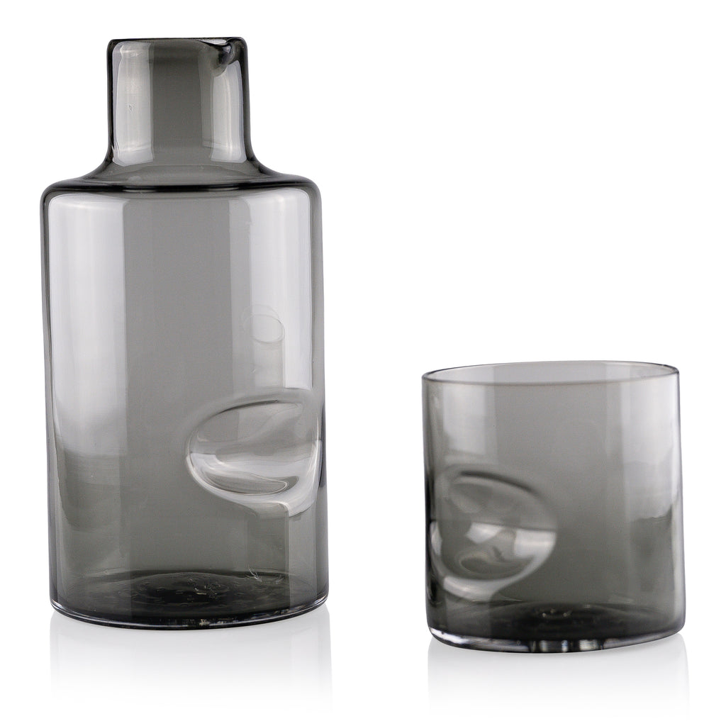 A sleek glass carafe and matching tumbler set on a reflective white surface, displaying minimalist design with smooth contours and subtle reflections, conveying a sense of modern elegance