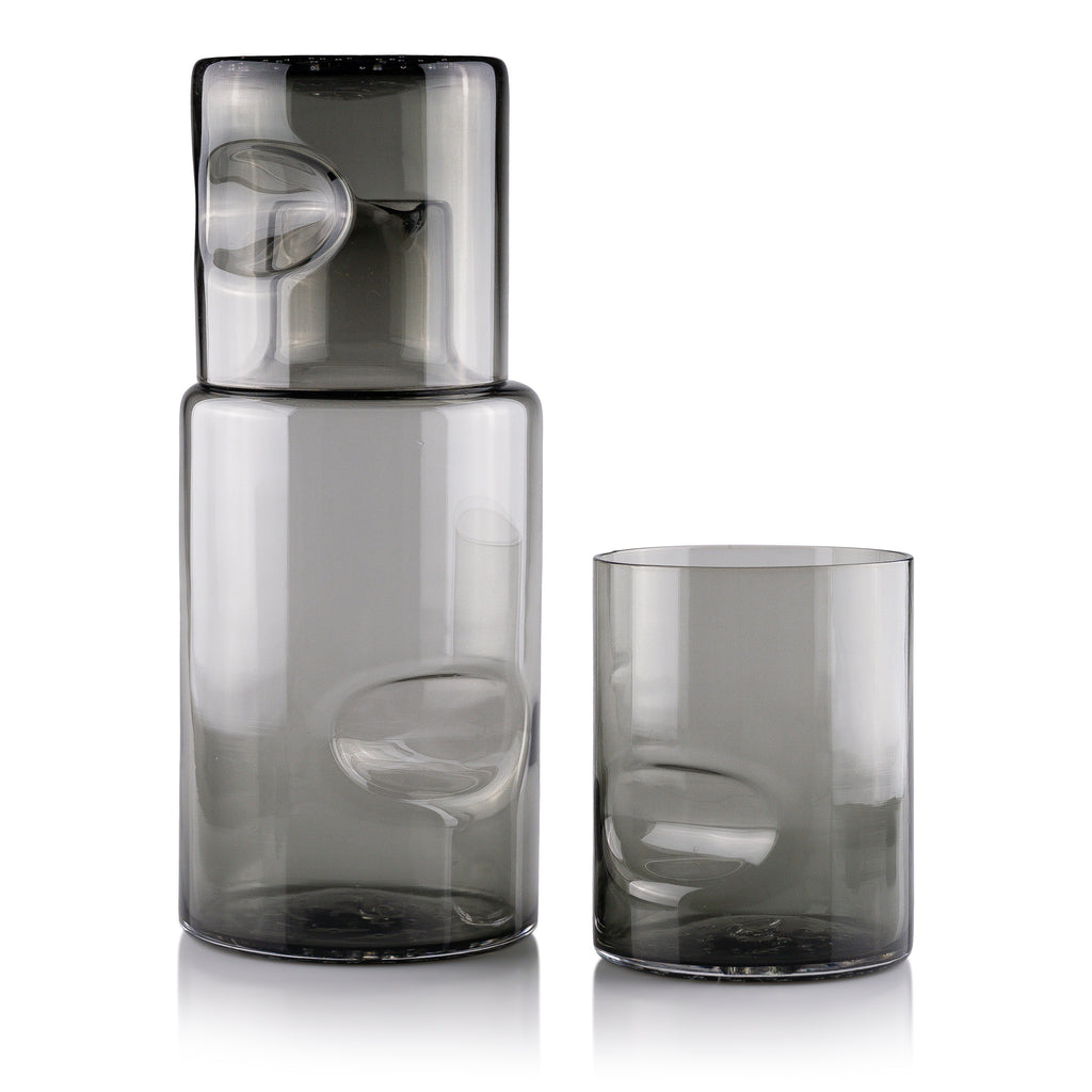 A sleek glass carafe and matching tumbler set on a reflective white surface, displaying minimalist design with smooth contours and subtle reflections, conveying a sense of modern elegance