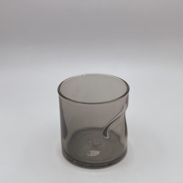 A contemporary smoky gray glass tumbler with a practical thumb indent for a secure grip. The glass's translucent quality allows light to filter through, softening its silhouette, and it's showcased against a white background that accentuates its modern and chic design.
