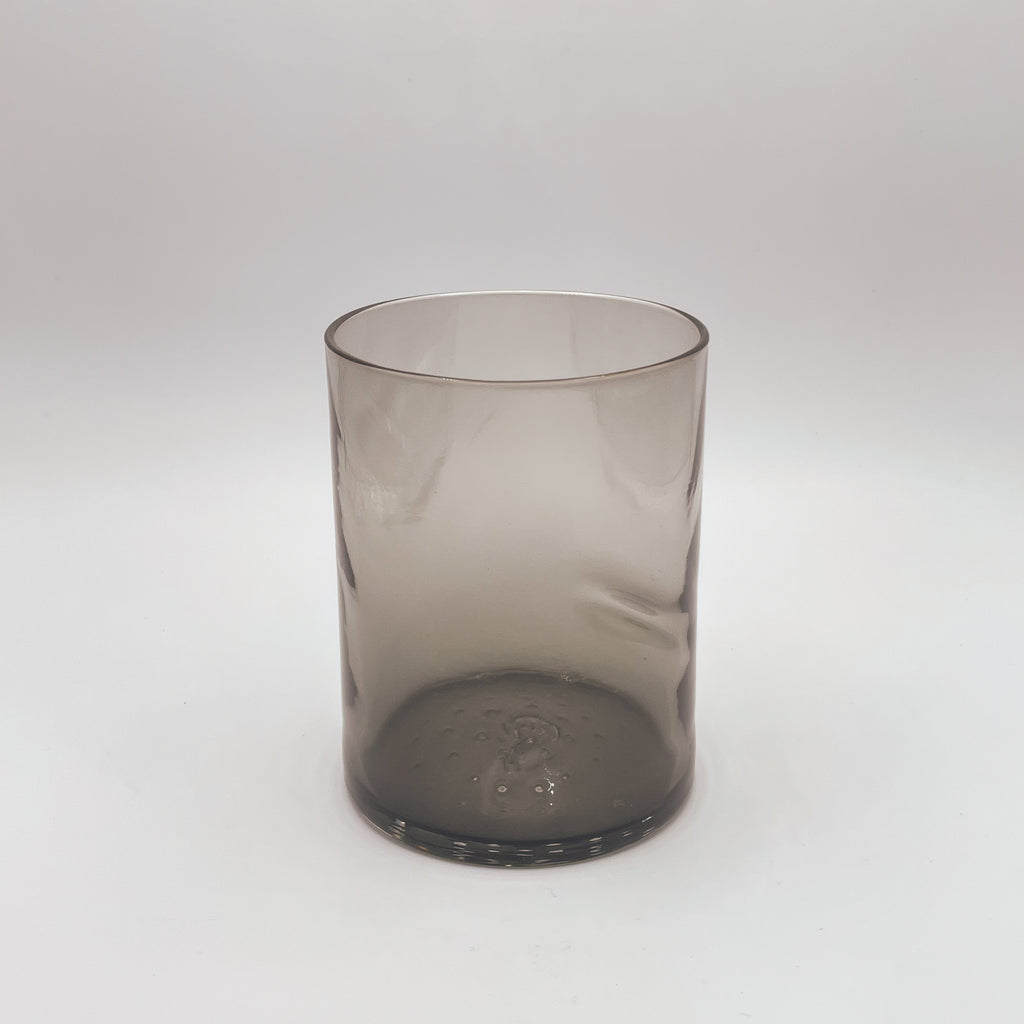 A contemporary glass tumbler with a smoky gradient, from transparent at the top to a deep charcoal at the base. It features a unique thumb indent, creating an ergonomic design. This glass is showcased against a white backdrop, which highlights its modern and stylish appearance.