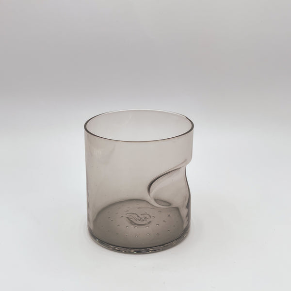 A modern smoky gray glass tumbler with a thumb indent design, set on a subtly textured white surface, evoking a minimalist and functional aesthetic.