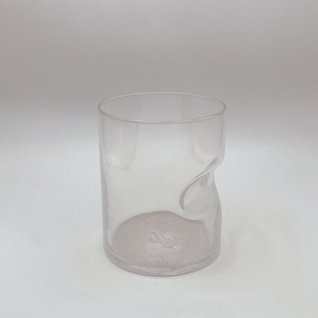 This image displays a clear glass tumbler with a unique thumb indent for ergonomic holding, completed with a subtle gold rim for an added touch of sophistication. The glass is placed against a white background, which emphasizes its transparency and clean design.