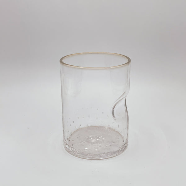 A transparent glass tumbler with a gracefully designed thumb indent, accentuated with a slight hint of color on the rim, creating a sophisticated simplicity, all set against a white backdrop that emphasizes its clean and clear appearance.