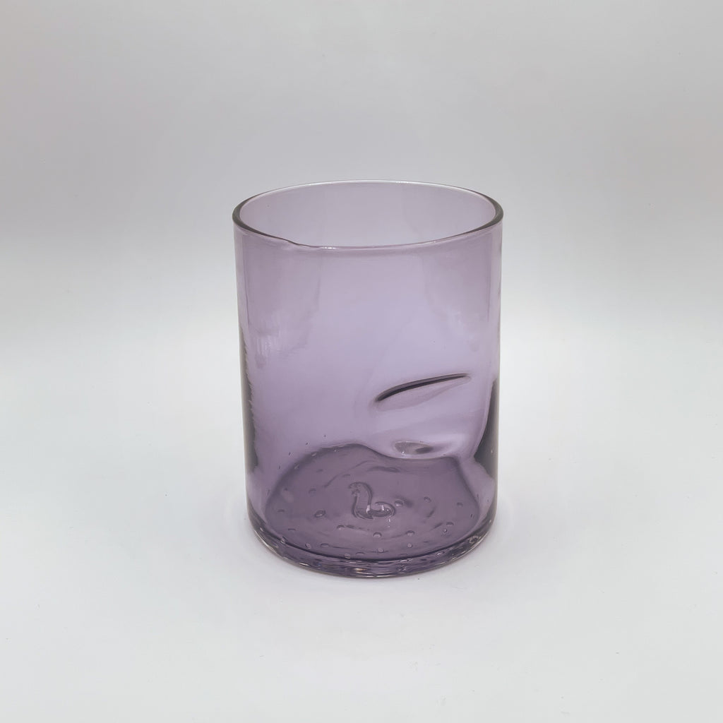A muted lavender glass tumbler features a thumb groove for ease of hold, showcasing a sophisticated gradient from a translucent top to a deeper hue at the base. Set against a white backdrop, the tumbler's simple lines and gentle color convey a contemporary elegance.