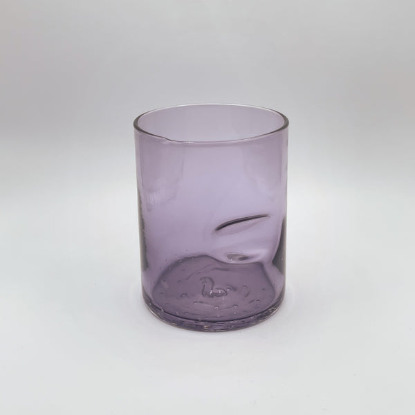 A muted lavender glass tumbler features a thumb groove for ease of hold, showcasing a sophisticated gradient from a translucent top to a deeper hue at the base. Set against a white backdrop, the tumbler's simple lines and gentle color convey a contemporary elegance.