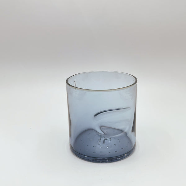 A handcrafted glass tumbler with a smokey blue gradient and a unique, swirled indentation at the side, adding an artistic touch. The tumbler has a thick base with small bubbles captured within the glass, reflecting meticulous craftsmanship, showcased against a soft white backdrop