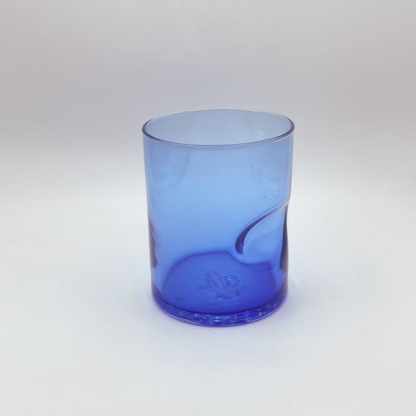 A vibrant cobalt blue glass tumbler with a convenient thumb indent, boasting a gradient that fades to a lighter blue at the top. This glass piece provides a bold pop of color, presented against a soft white background that subtly highlights the gradient effect.
