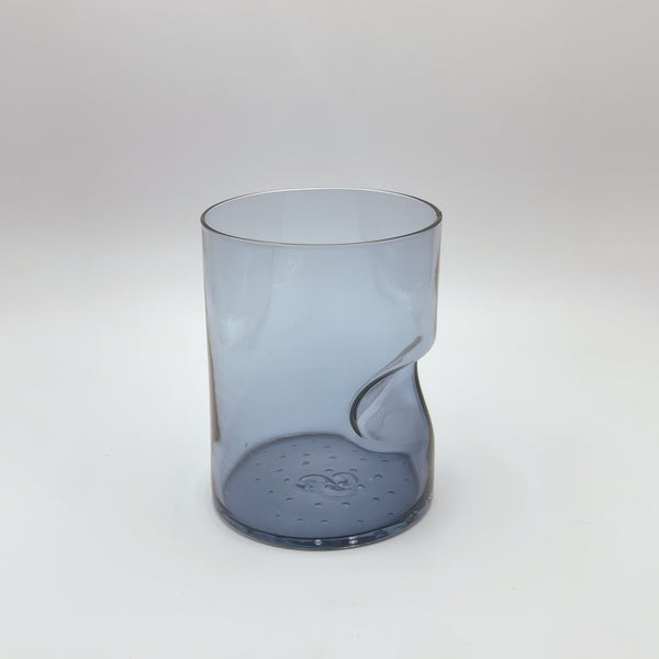 A chic blue glass tumbler with a thumb indent for easy gripping, exhibiting a subtle color and translucent quality. The tumbler's soft, muted blue tone fades into clarity towards its rim, showcased on a white background that accentuates its sleek design.