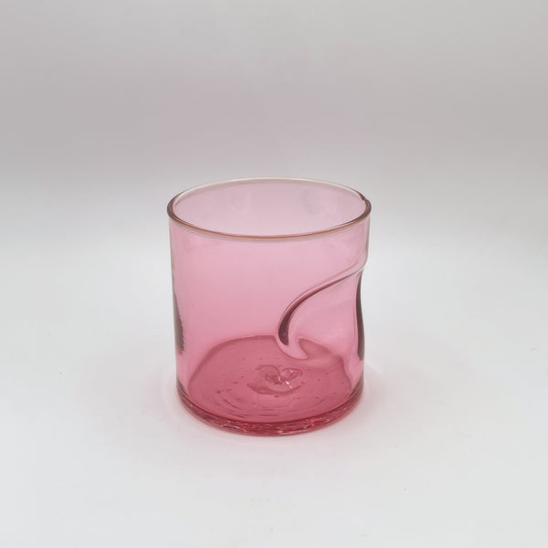 A charming glass tumbler tinted in a gradient of pink, from a transparent top to a more concentrated hue at the base, complete with a comfortable thumb indent, photographed against a gentle white background, which subtly complements its soft color.