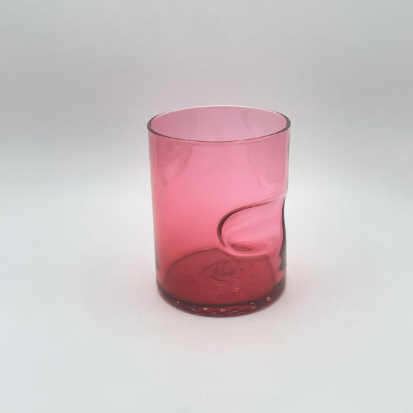 This image features a vibrant glass tumbler in a rich shade of fuchsia, fading to clear towards the top. The glass showcases a unique thumb indent, adding to its modern appeal. It's set against a soft, white background that accentuates the striking color gradient.