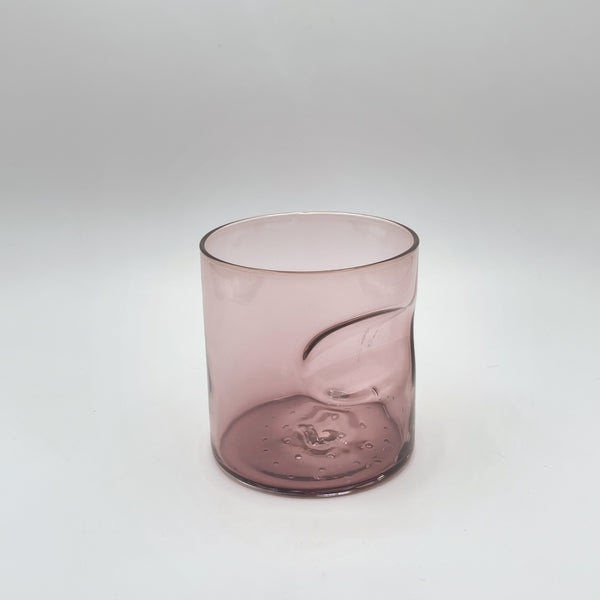A delicate blush pink glass tumbler with a thumb indent detail, offering a touch of elegance with its simple yet sophisticated design, set against a soft white background that underscores its subtle hue and clean lines.