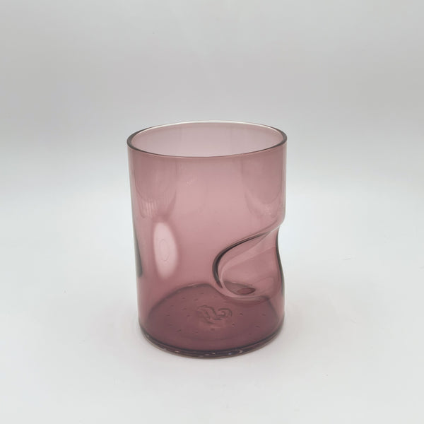 A soft mauve glass tumbler, enhanced with a thoughtful thumb indent, displays a gentle gradient from deeper color at the base to a light translucence at the top. The tumbler stands against a white backdrop, emphasizing its subtle and elegant color variation.