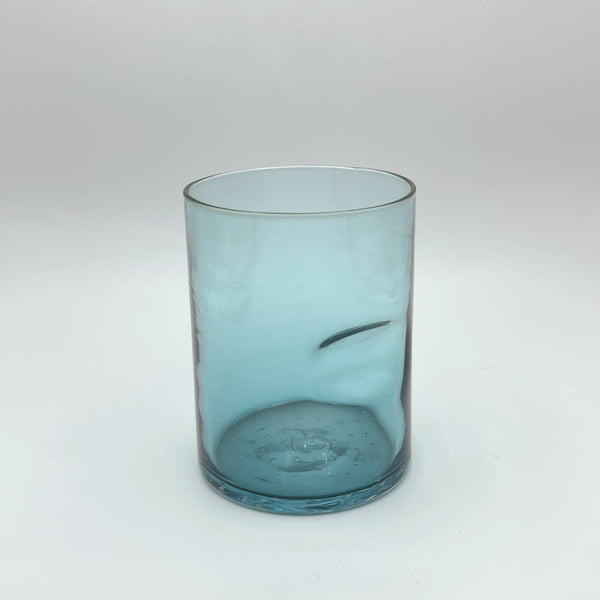 A delicate aqua blue glass tumbler positioned on a plain white background. The tumbler features an elegant swirl detail near its base, creating a visual effect reminiscent of gentle waves. The clear glass rim and the subtle texture on the bottom of the tumbler add to its artisanal charm.