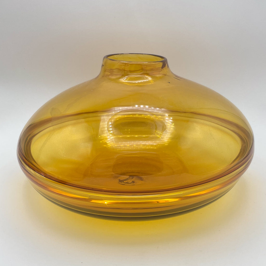 A wide, spherical amber glass vase with a flattened bottom and a short, narrow neck. The vase has a soft translucence, capturing and diffusing the light around it, showcasing the intricate details and subtle variations in color. The design includes a concentric ring pattern on the base, which adds to its handcrafted appeal against a gradient white to warm background.