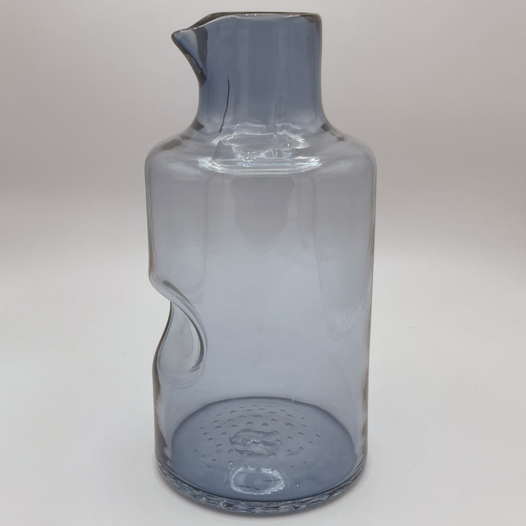 A sleek, smoky blue glass carafe with an elegant spout and an innovative thumb indent for easy pouring. Its contemporary design features a slender, slightly curved body, set against a white background to highlight its modern, functional aesthetic.