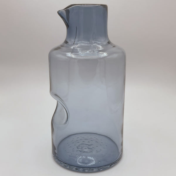 A sleek, smoky blue glass carafe with an elegant spout and an innovative thumb indent for easy pouring. Its contemporary design features a slender, slightly curved body, set against a white background to highlight its modern, functional aesthetic.