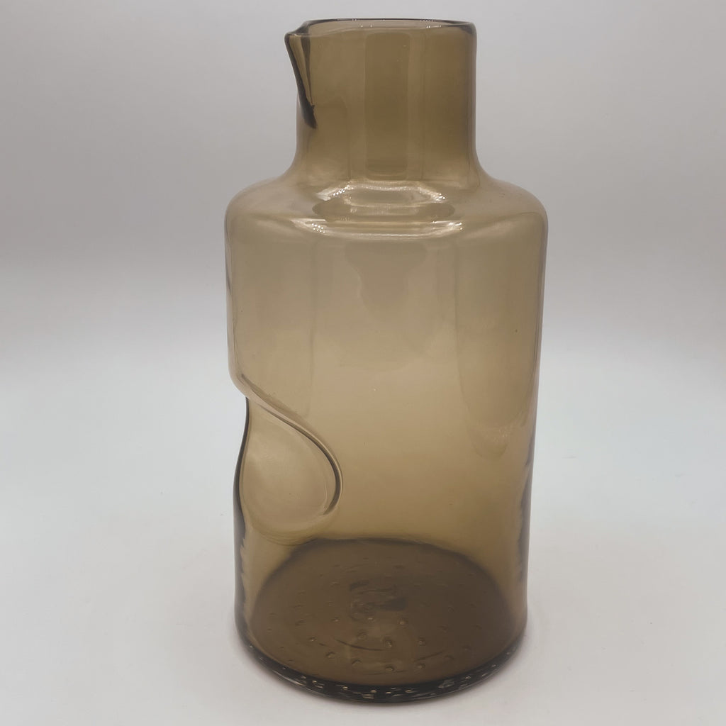 A tall, amber-hued glass carafe with a distinctive modern design featuring a smooth, elongated neck and an asymmetrical indented curve on its body, enhancing its visual appeal. The carafe stands on a dark-textured base against a soft white background, highlighting its transparent and reflective surface.