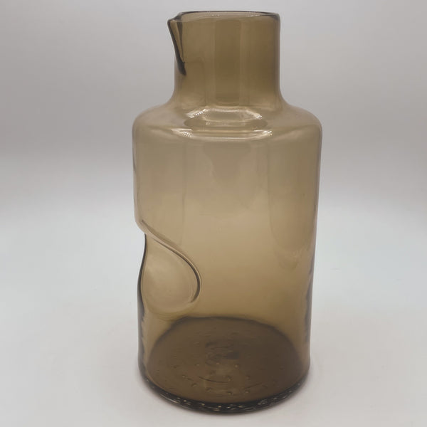 A tall, amber-hued glass carafe with a distinctive modern design featuring a smooth, elongated neck and an asymmetrical indented curve on its body, enhancing its visual appeal. The carafe stands on a dark-textured base against a soft white background, highlighting its transparent and reflective surface.