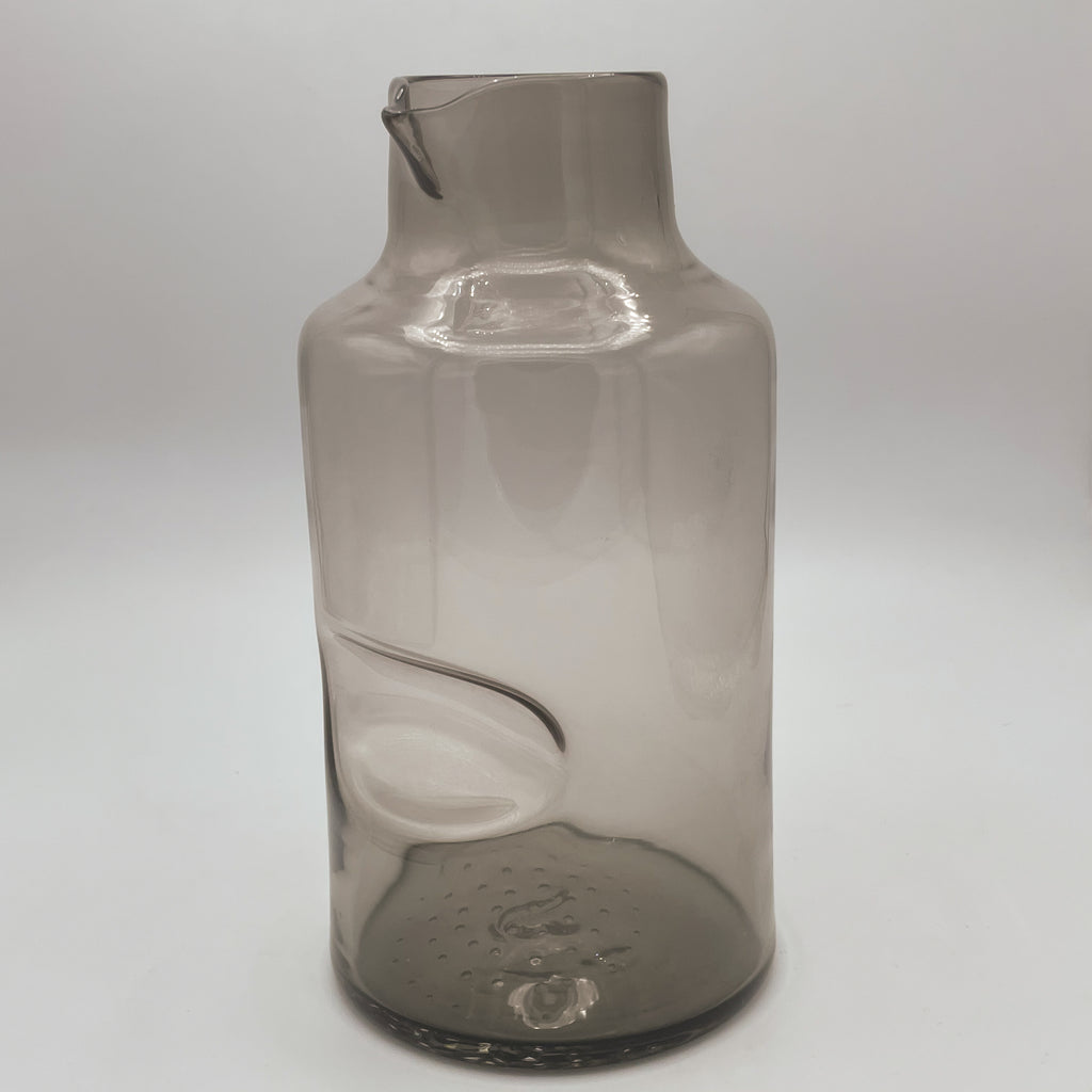 A smoky gray glass carafe with a stylish thumb indent and a smartly designed spout for easy pouring. The carafe's tall and slender shape gives it a modern look, perfect for contemporary table settings, displayed against a light background that highlights its sleek design and translucent quality.