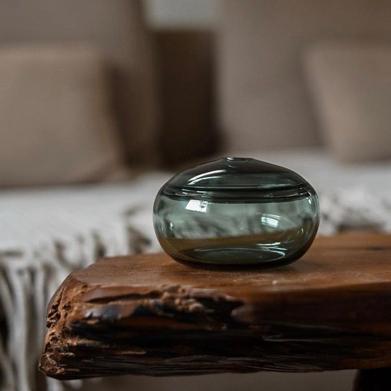 A squat glass vessel with a wide, flattened body and a narrow, raised lid, photographed against a white background, showcasing its smooth lines and tranquil, translucent color, reflective of serene, contemporary glass artistry.